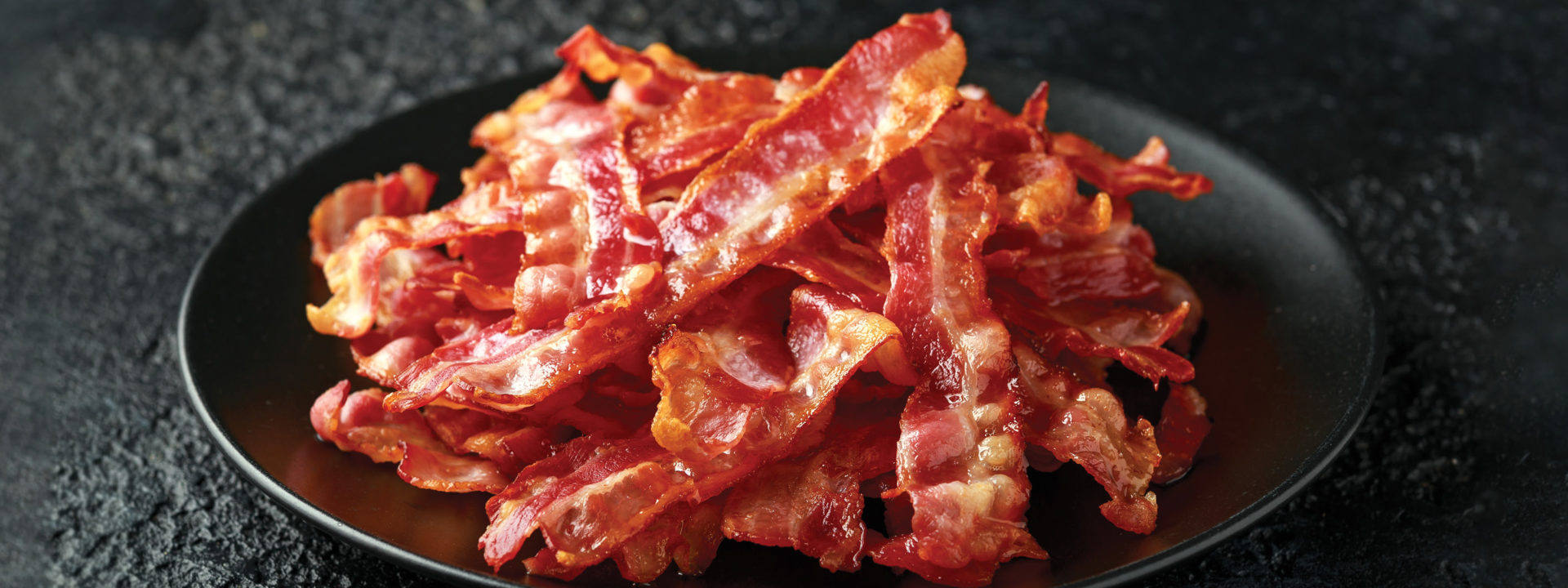 Stack of cooked bacon on a black plate on a black concrete countertop