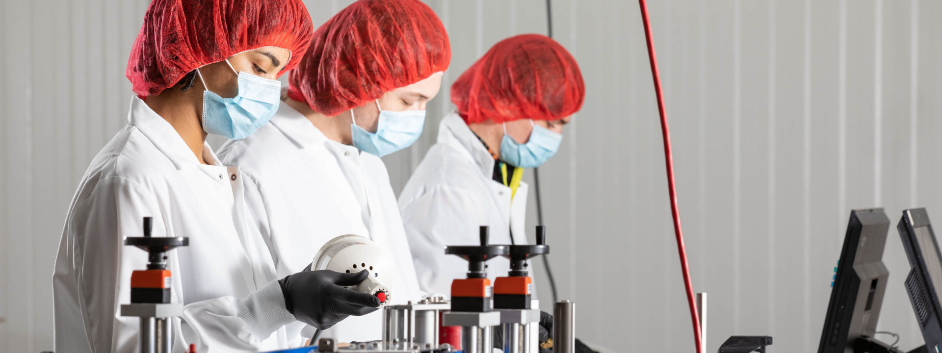 Three Indiva employees with protective work gear including white coats, black gloves and face masks working in the Production area with manufacturing equipment.