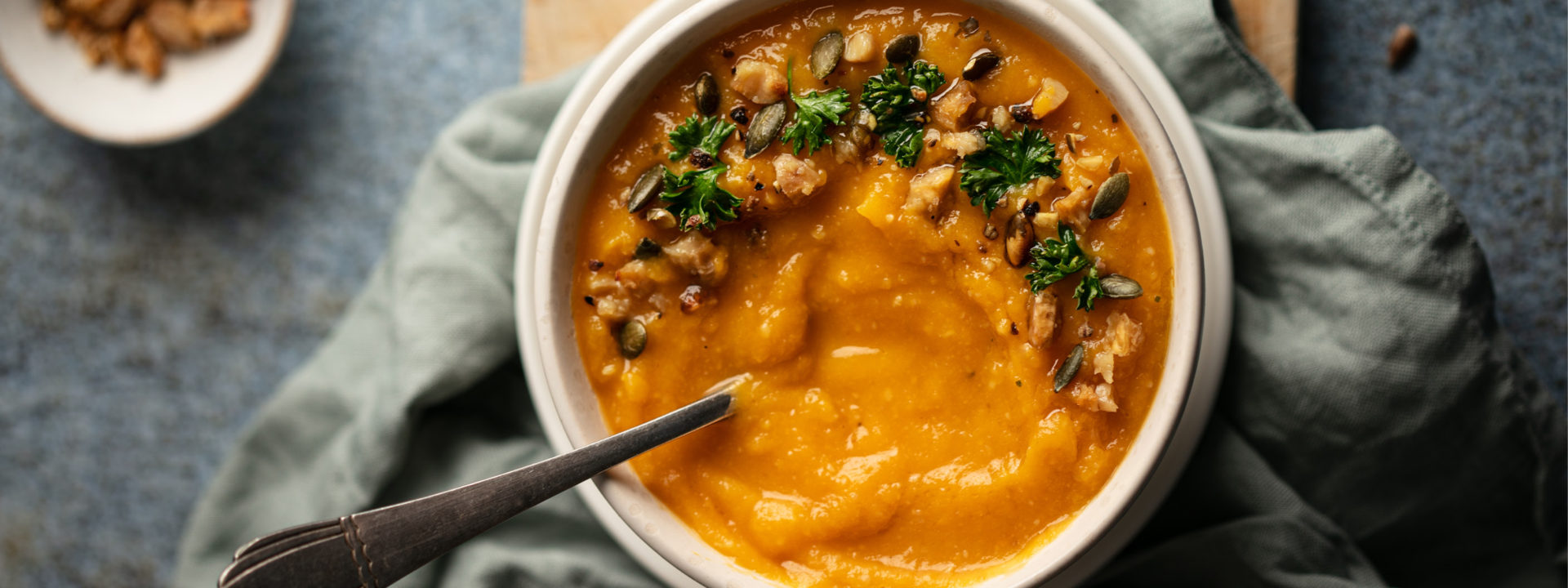 Overhead shot of bowl filled with chestnut and squash soup with pieces of parsley on top and spoon in bowl. Bowl placed on a wooden charcuterie board with a grey linen napkin and bowl of chestnuts beside