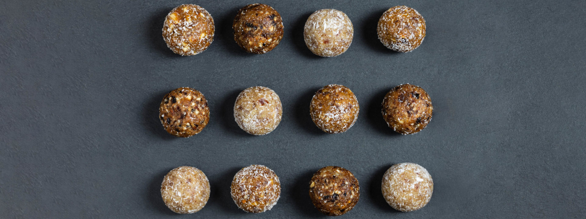 Flat lay 12 symmetrical mixed flavour energy balls arranged lined up on a charcoal grey background