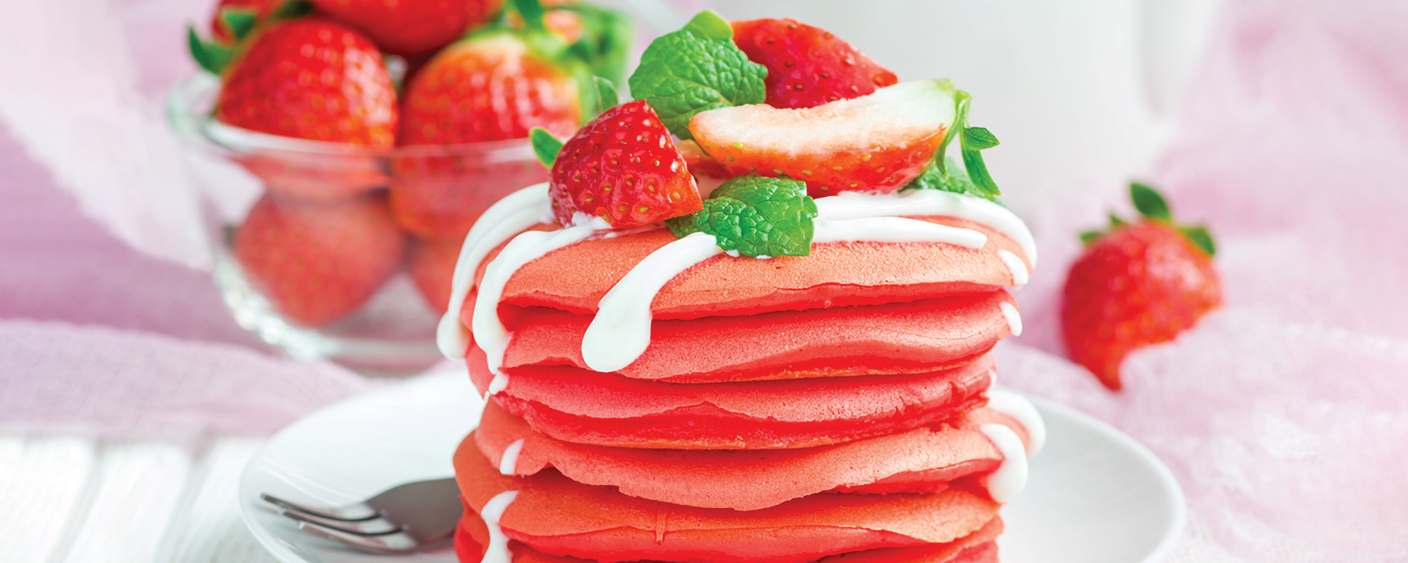 Stack fluffy red pancakes with strawberries, mint and whipped cream on top, on a white plate with a silver fork beside
