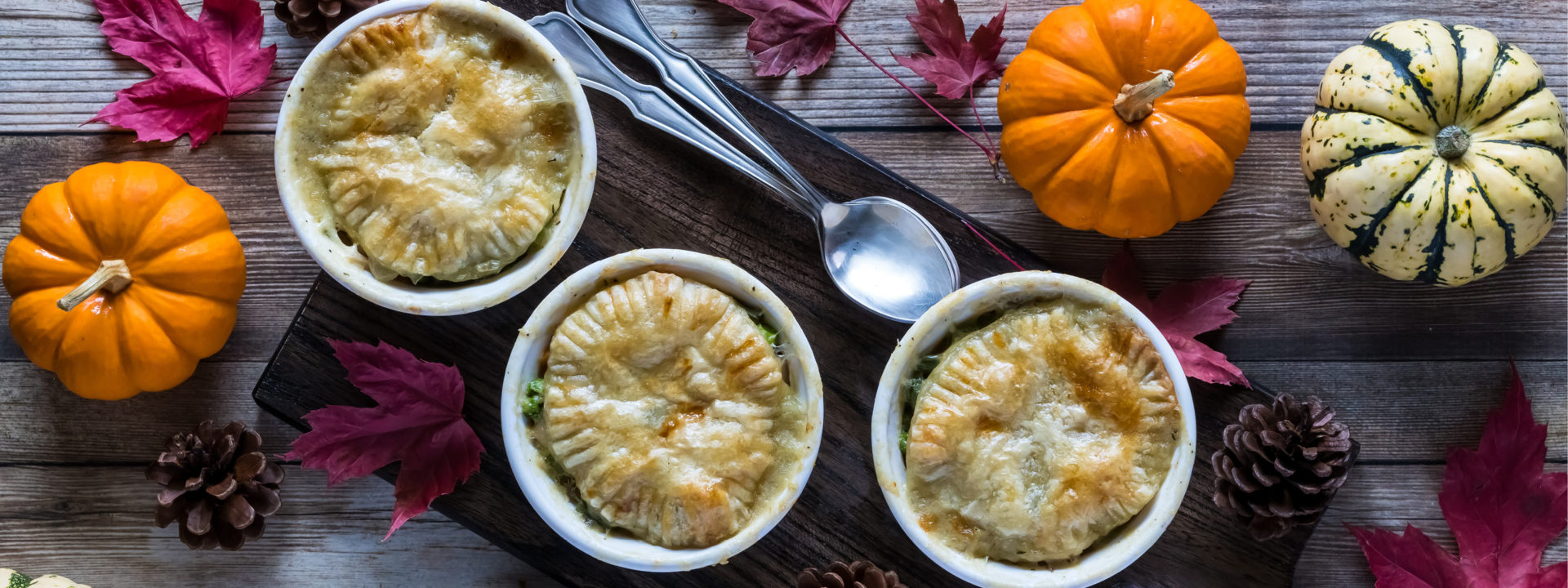 Three small turkey pot pies on a wooden plant serving board, seen from above with two spoons, pinecones, pumpkins and leaves around them